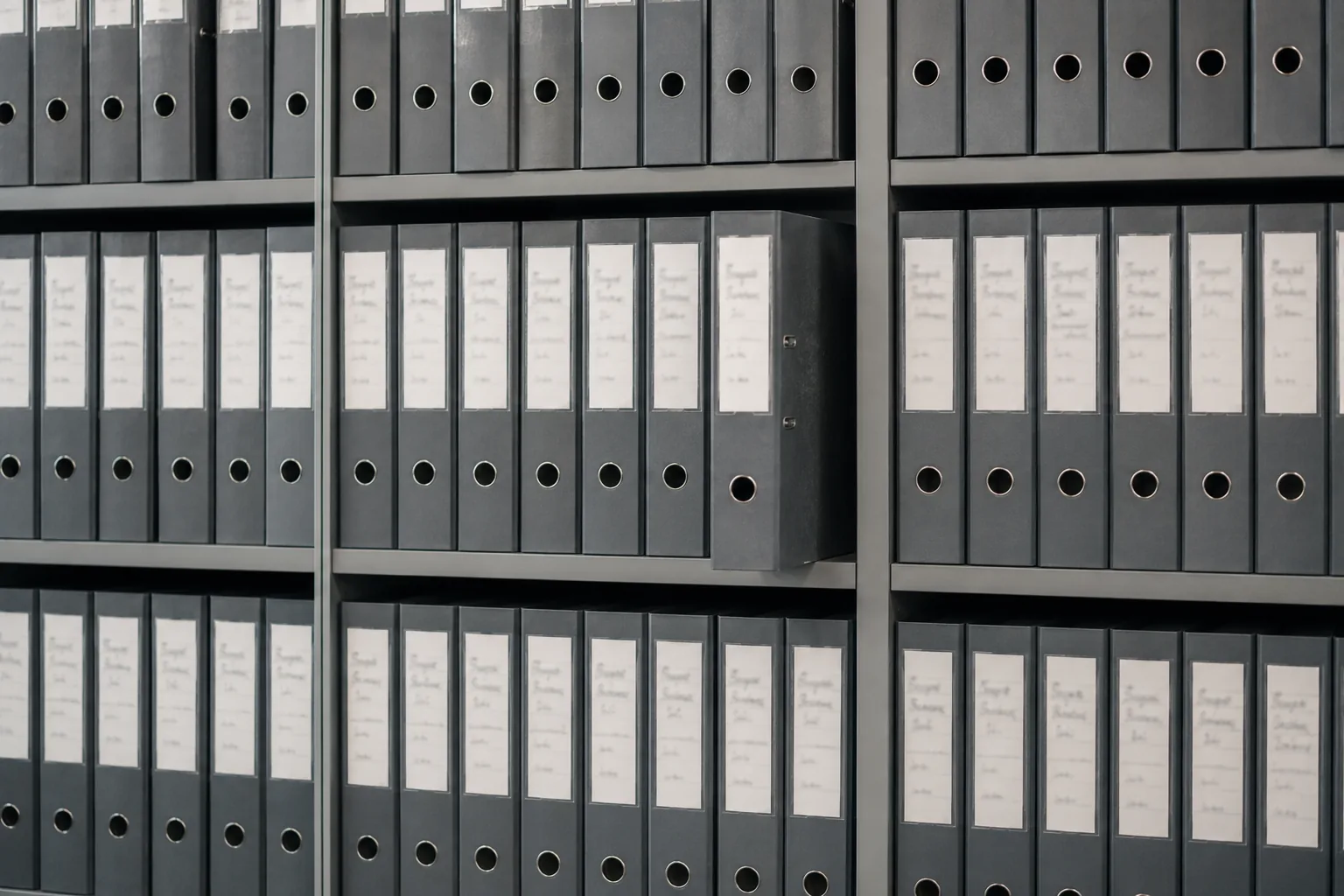 Regalwand mit gleichförmigen dunkelgrauen Aktenordnern, ein Ordner steht vor.